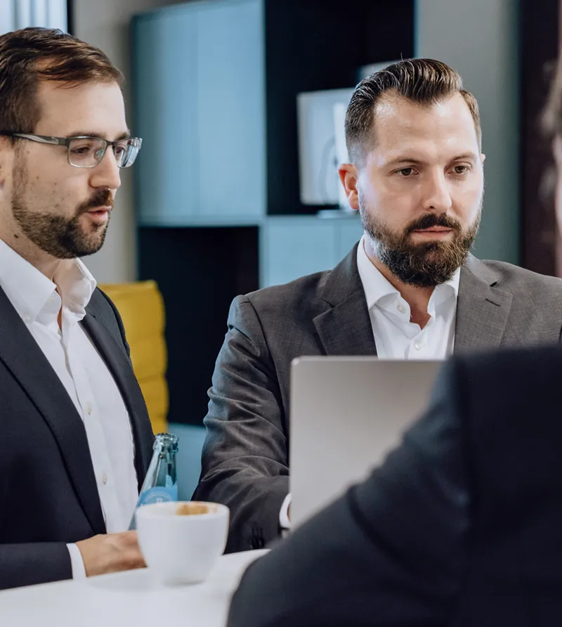 Three people in business attire having a discussion around a laptop at a white table, with coffee and water bottles present.