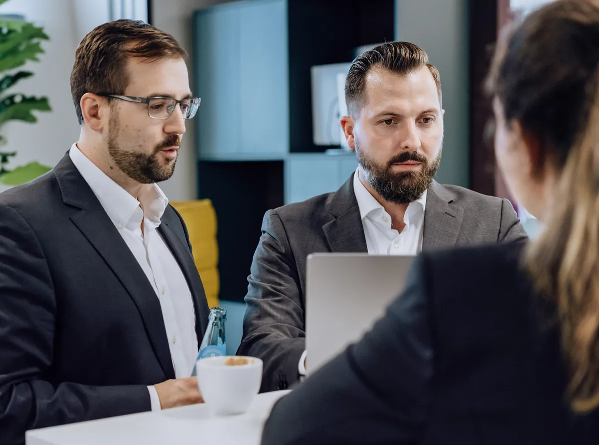 Three people in business attire having a discussion around a laptop at a white table, with coffee and water bottles present.