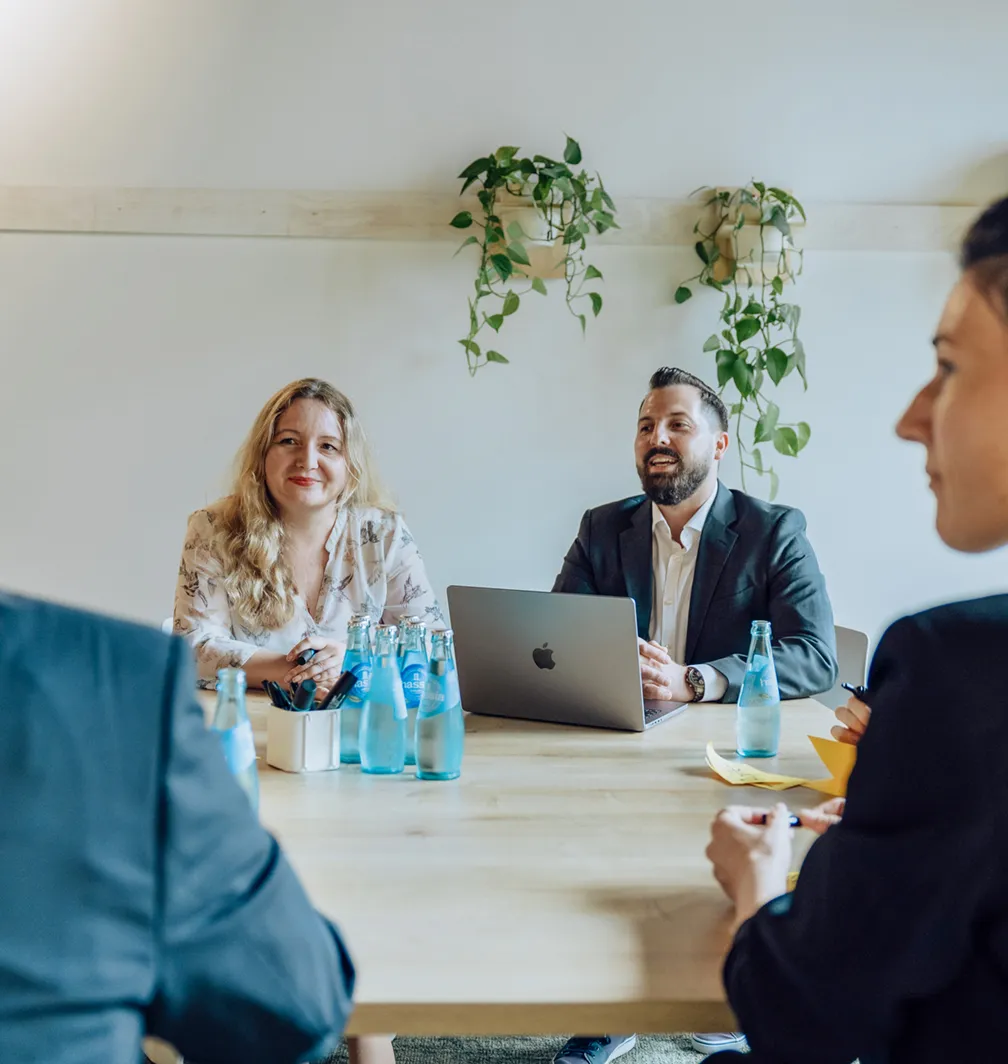 A group of four people in a business meeting, sitting around a table with laptops and water bottles, in a room with plants on the wall.