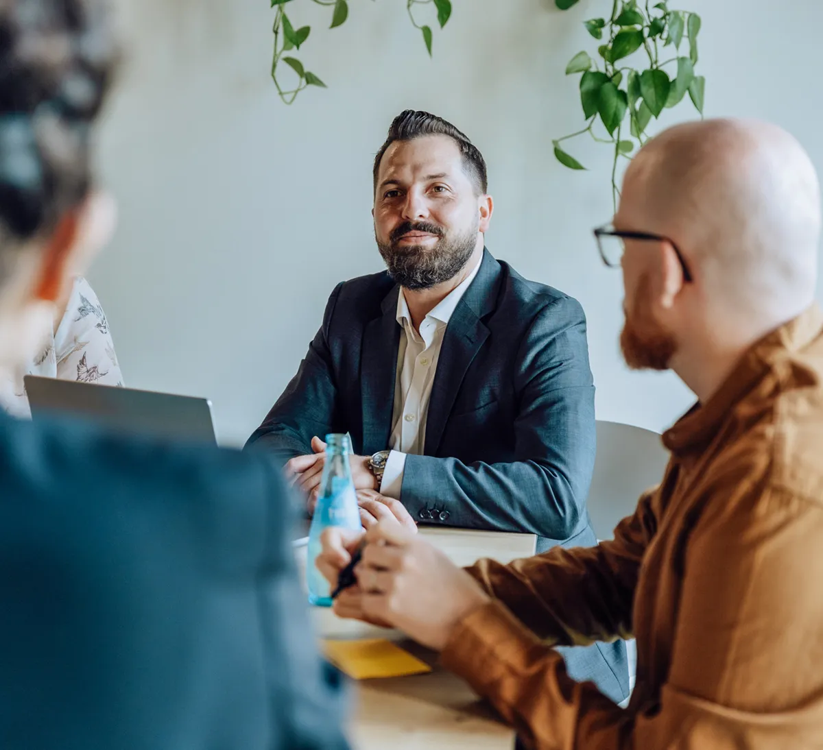 A group of people in a meeting room, with a man in a suit smiling and others listening attentively. There are laptops and a bottle on the table, and plants hanging in the background.