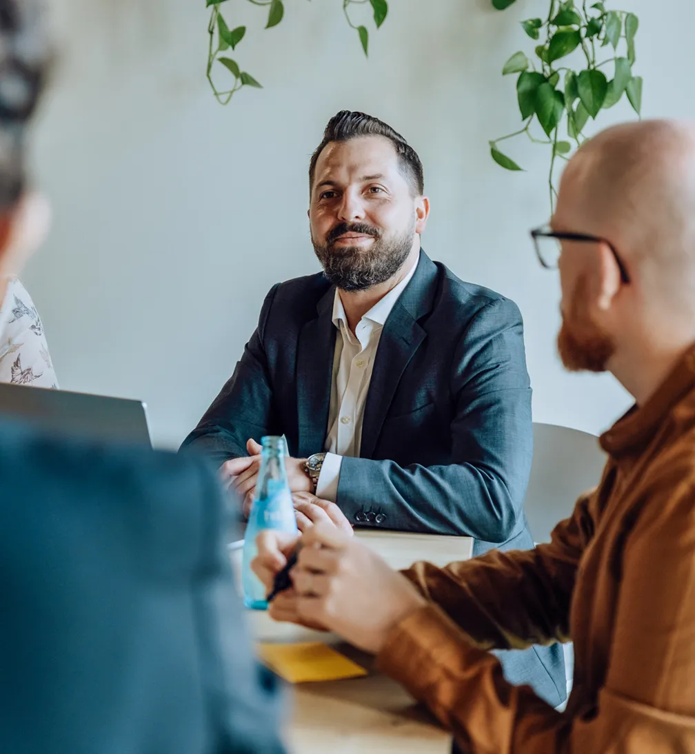 A group of people in a meeting room, with a man in a suit smiling and others listening attentively. There are laptops and a bottle on the table, and plants hanging in the background.