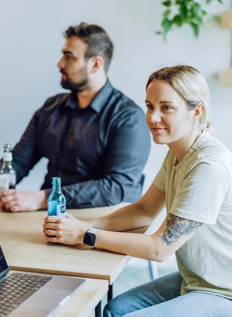 A group of four people sitting around a table with laptops and drinks, engaged in a casual meeting. One person is holding a blue bottle, and another has a laptop with a sticker that reads 'GIRL BOSS'.