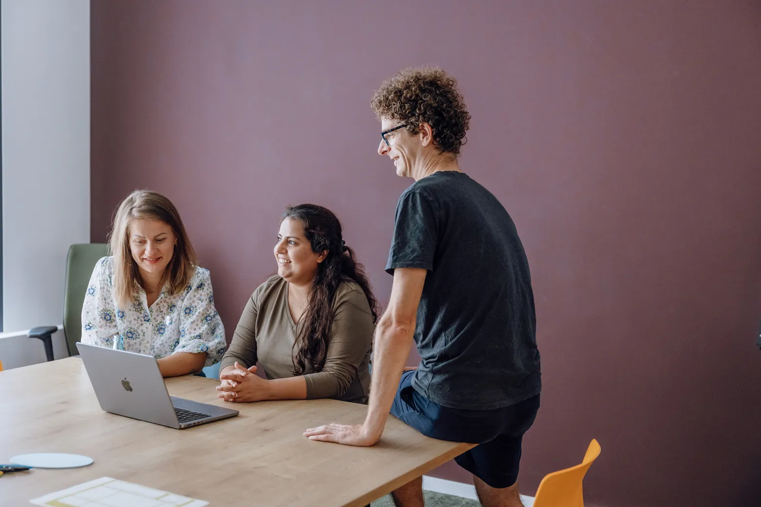 Three people having a casual meeting around a table with a laptop, smiling and engaged in conversation.
