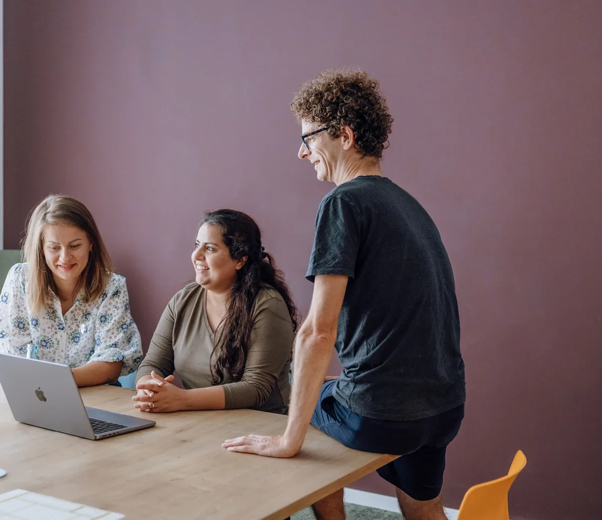 Three people having a casual meeting around a table with a laptop, smiling and engaged in conversation.