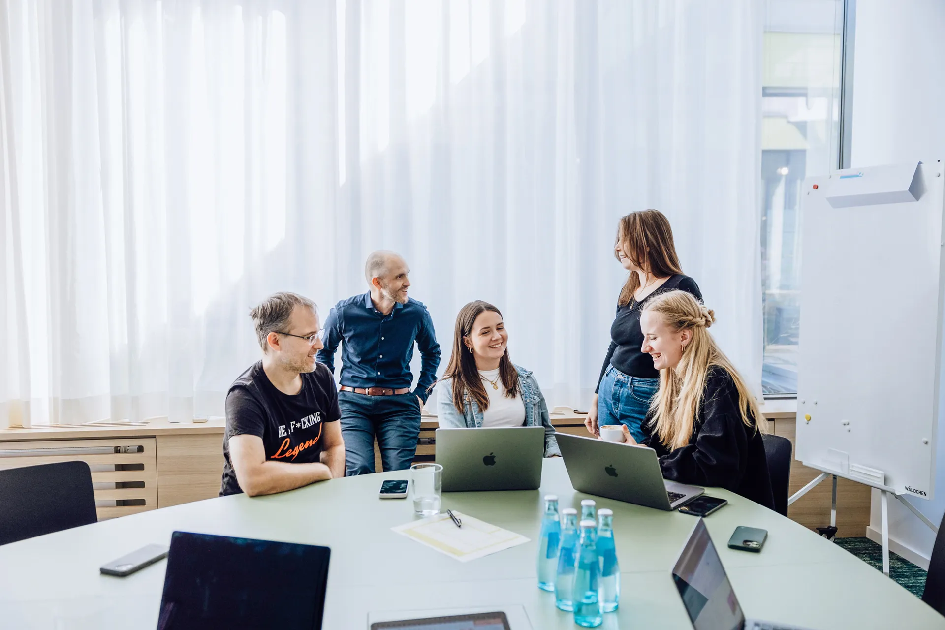 A group of five people having a casual meeting around a table with laptops and water bottles, in a bright room with large windows and a whiteboard.