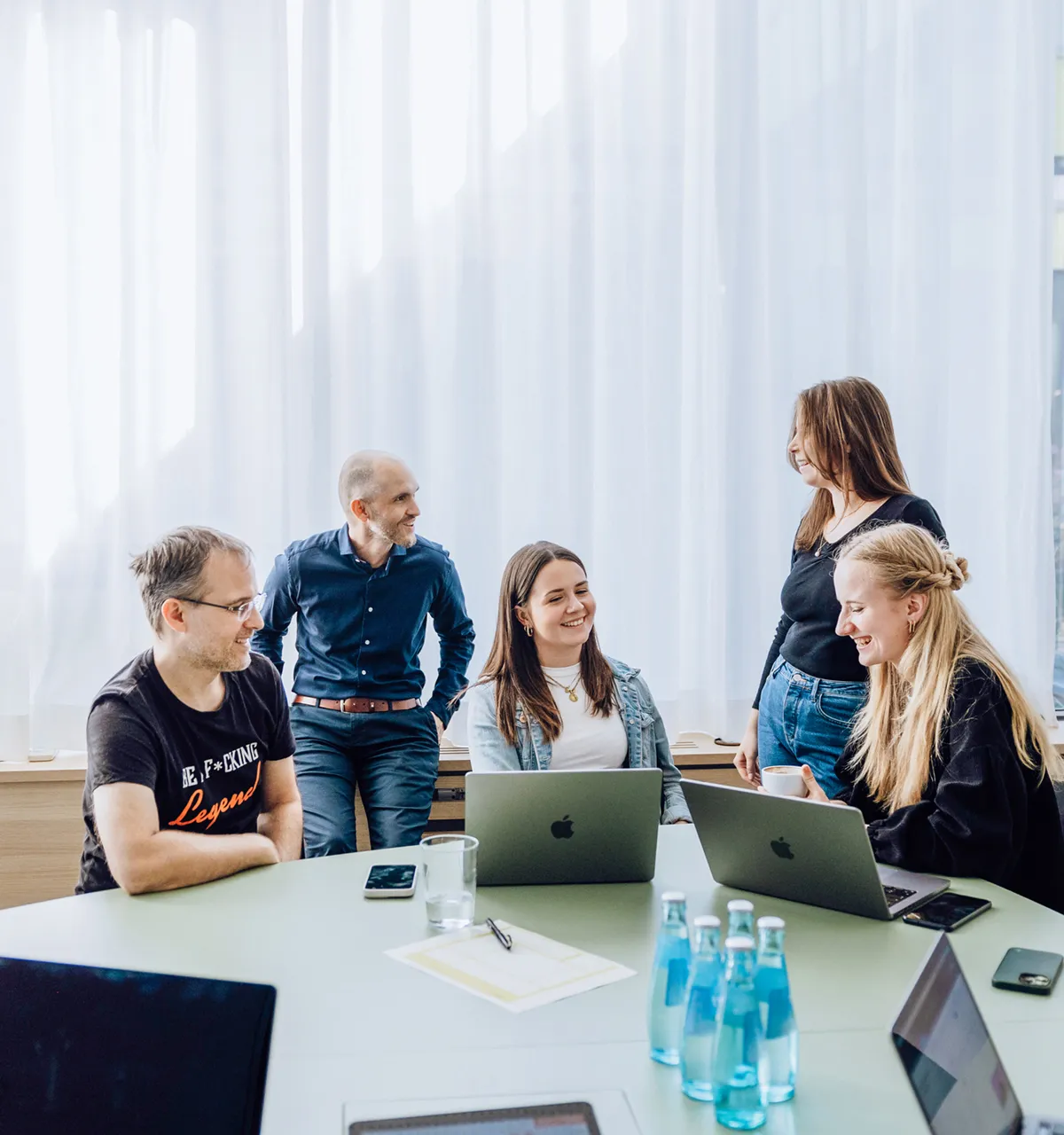 A group of five people having a casual meeting around a table with laptops and water bottles, in a bright room with large windows and a whiteboard.
