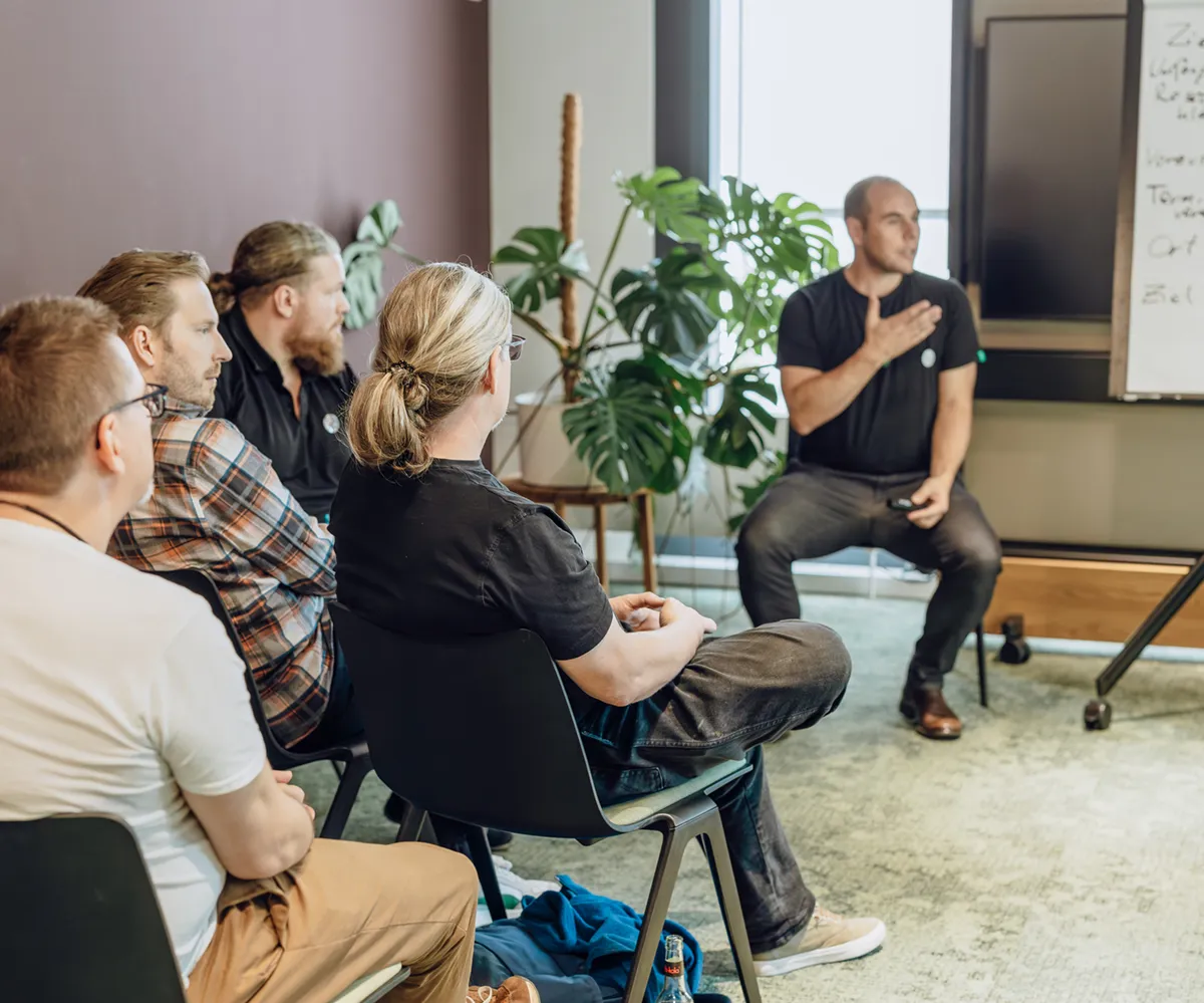 A group of people sitting in chairs attentively listening to a speaker in a casual office setting. The speaker is gesturing towards a flip chart with notes. There are plants in the background.