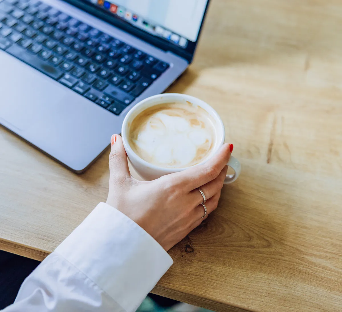 A person holding a cup of coffee next to an open laptop on a wooden table.