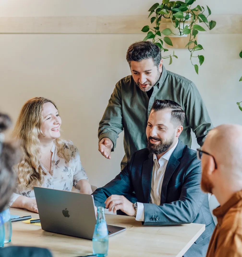 Group of colleagues having a discussion around a table with a laptop and water bottles, surrounded by hanging plants.
