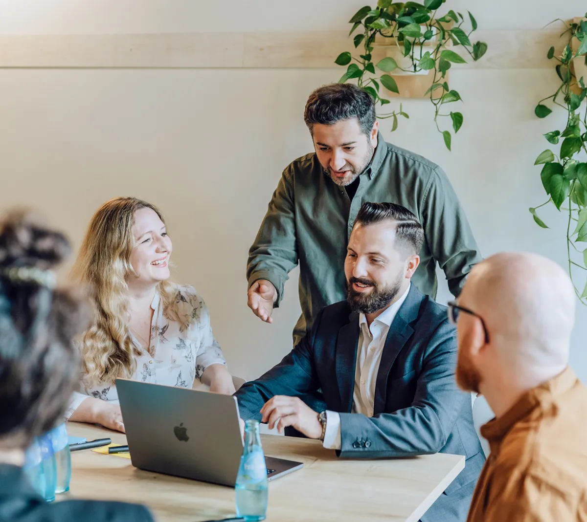 Group of colleagues having a discussion around a table with a laptop and water bottles, surrounded by hanging plants.