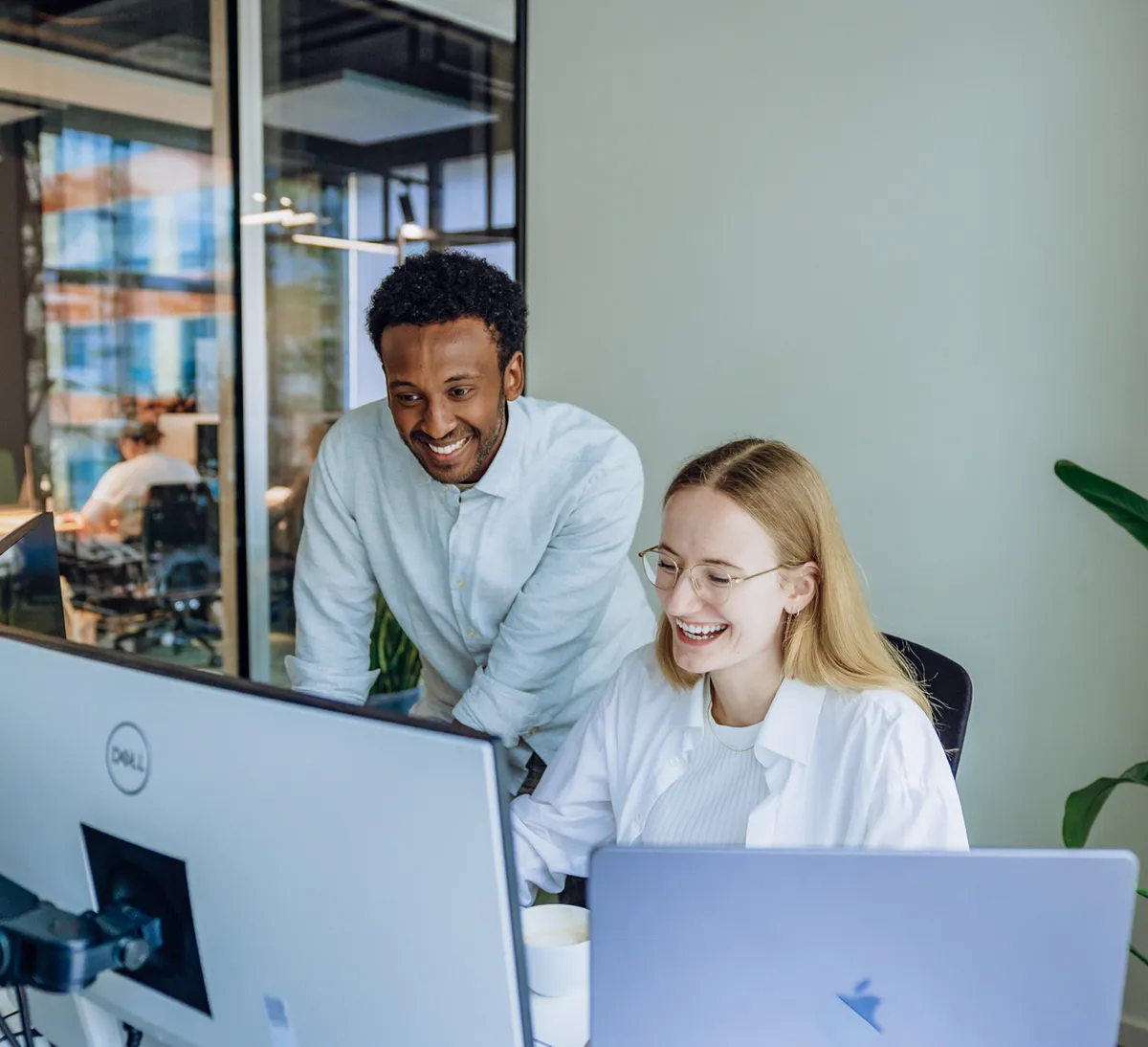 Two colleagues smiling and working together at a desk with computers in a modern office setting.