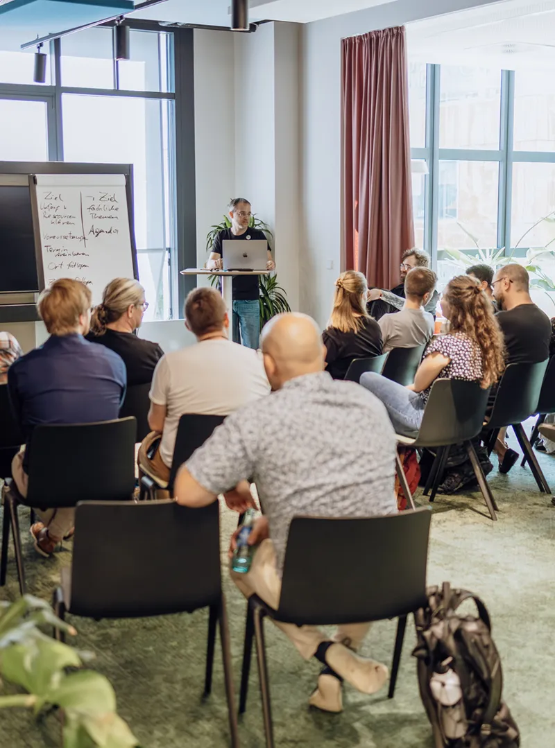 A group of people seated in a conference room, attentively listening to a speaker standing at the front with a laptop and a whiteboard displaying notes.