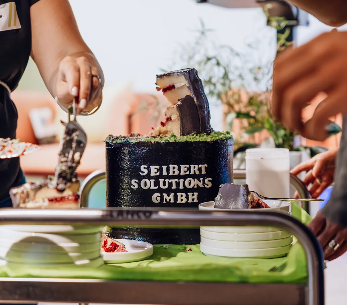 A person cutting a cake with the text 'Seibert Solutions GmbH' on it, placed on a table with plates and utensils.