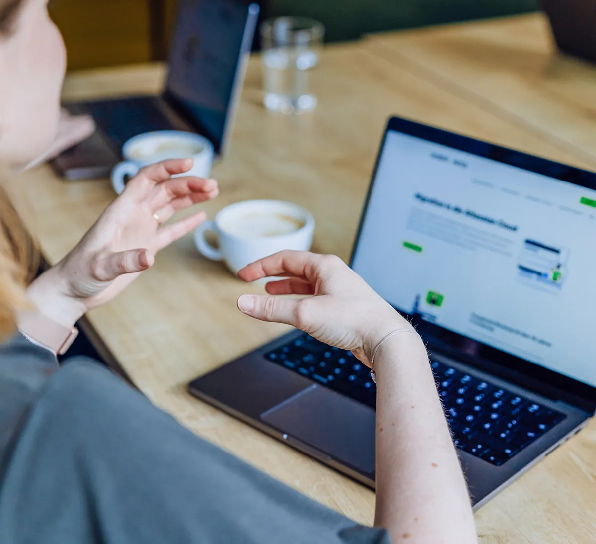 Person gesturing while discussing content on a laptop screen with coffee cups on the table.