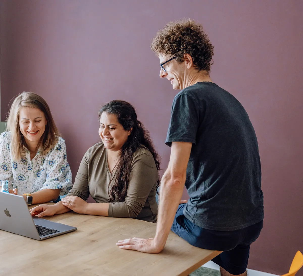 Three people gathered around a laptop on a wooden table, smiling and engaged in conversation.