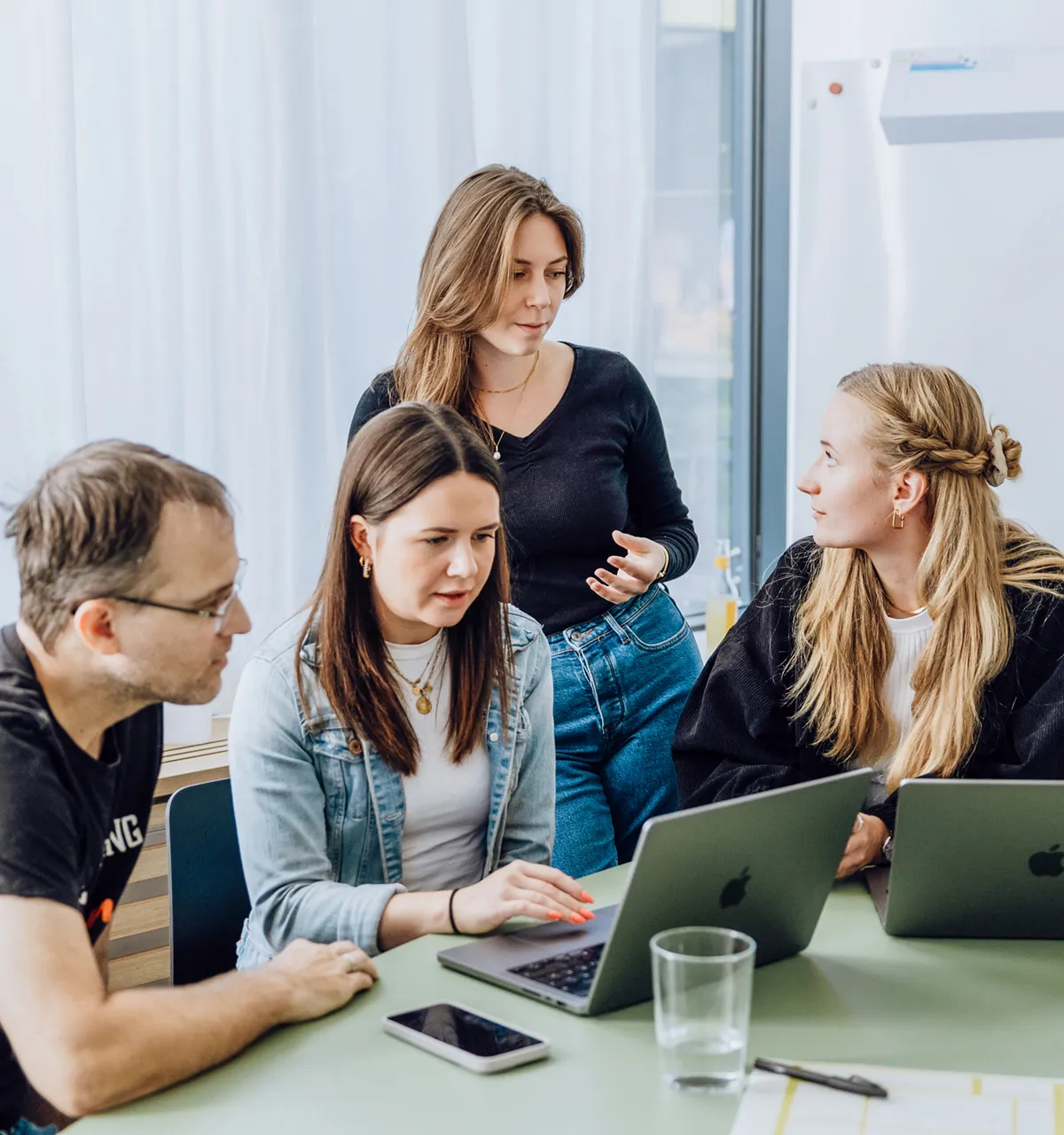 Four people gathered around a table with laptops, engaged in a discussion. One person is standing, while the others are seated, focusing on the laptops.