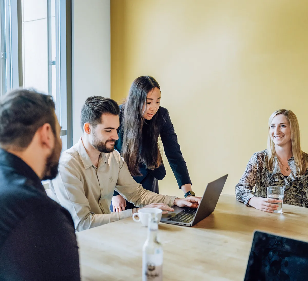 A group of people sitting around a table with laptops, engaged in a discussion. One person is standing and pointing at a laptop screen, while others are seated, smiling and holding drinks. The setting is a bright room with a yellow wall.