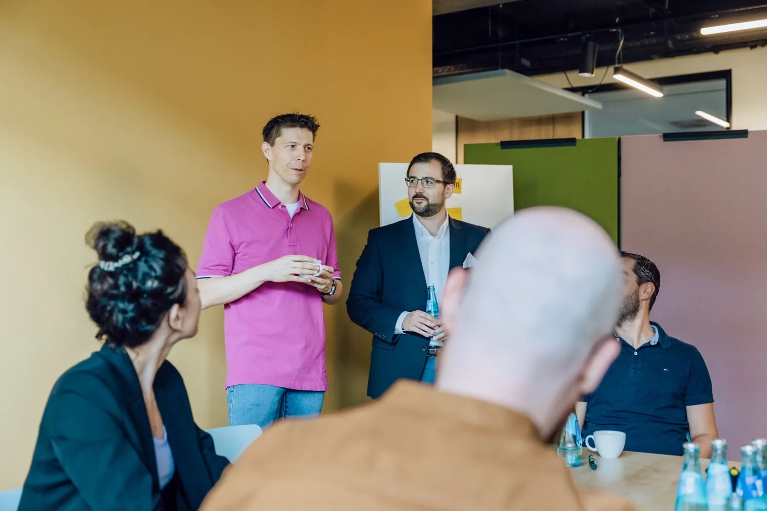A group of people in a meeting room with a yellow wall, engaged in conversation. One person in a pink shirt is speaking while others listen attentively.