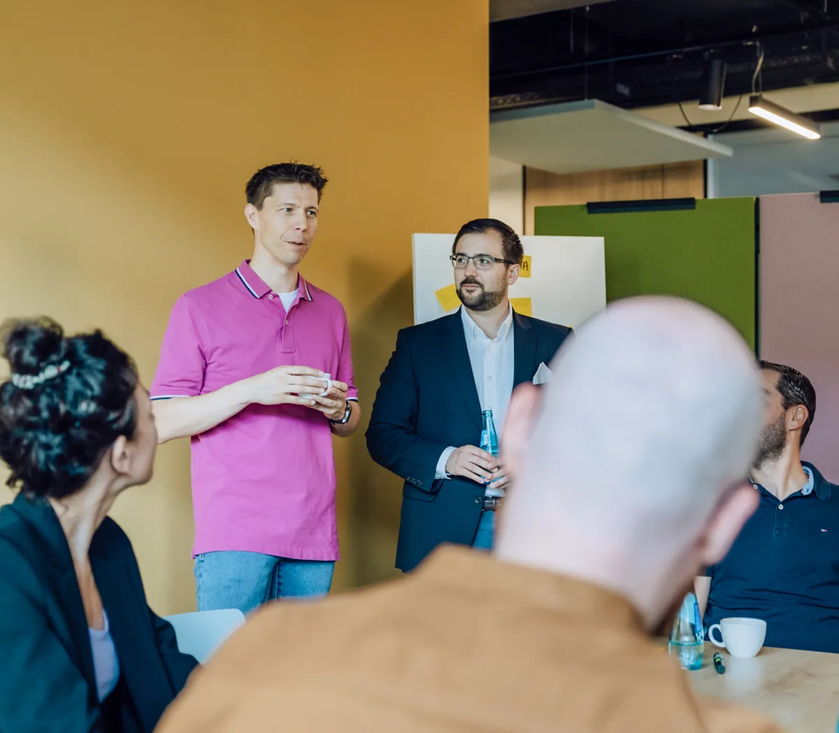 A group of people in a meeting room with a yellow wall, engaged in conversation. One person in a pink shirt is speaking while others listen attentively.