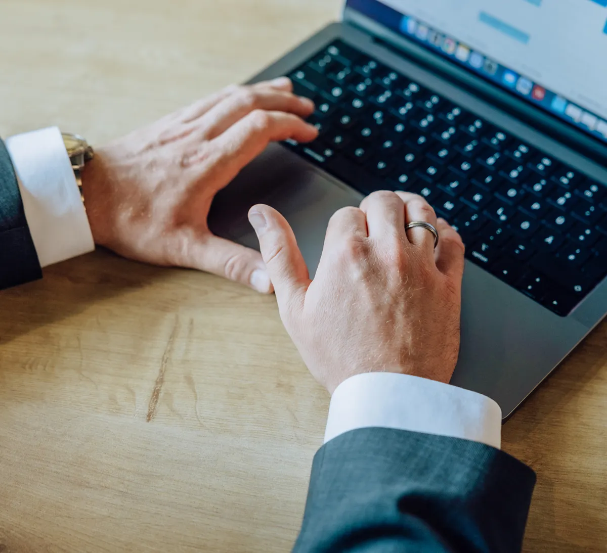 Close-up of hands typing on a laptop keyboard, wearing a suit and ring.