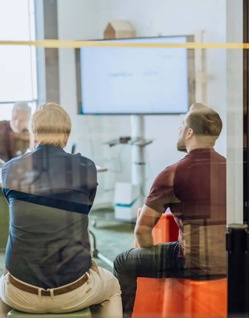 A group of people sitting in a modern office room, attentively watching a presentation on a large screen. The room is visible through a glass wall.