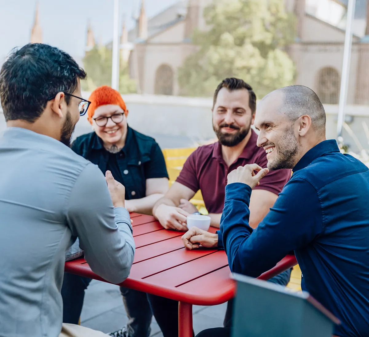A group of five people sitting around a red table outdoors, engaged in a lively conversation. They are smiling and appear to be enjoying each other's company. The background shows a blurred view of buildings and greenery.