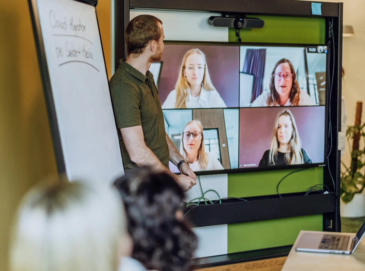 A man standing in front of a large screen displaying a video conference with four participants. A flipchart is visible on the left side of the image.