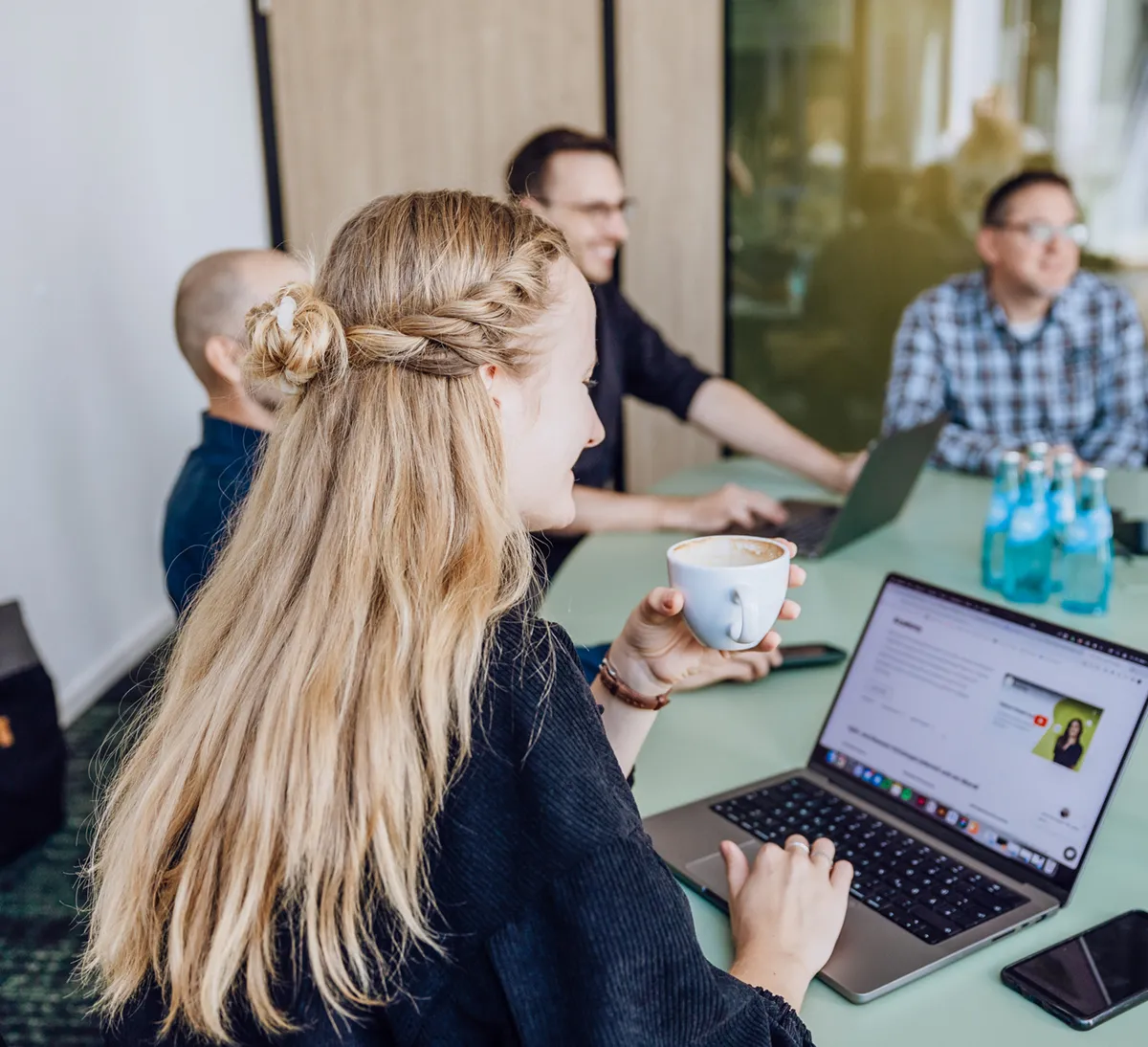 A woman with long blonde hair holding a coffee cup while using a laptop in a meeting room with colleagues.