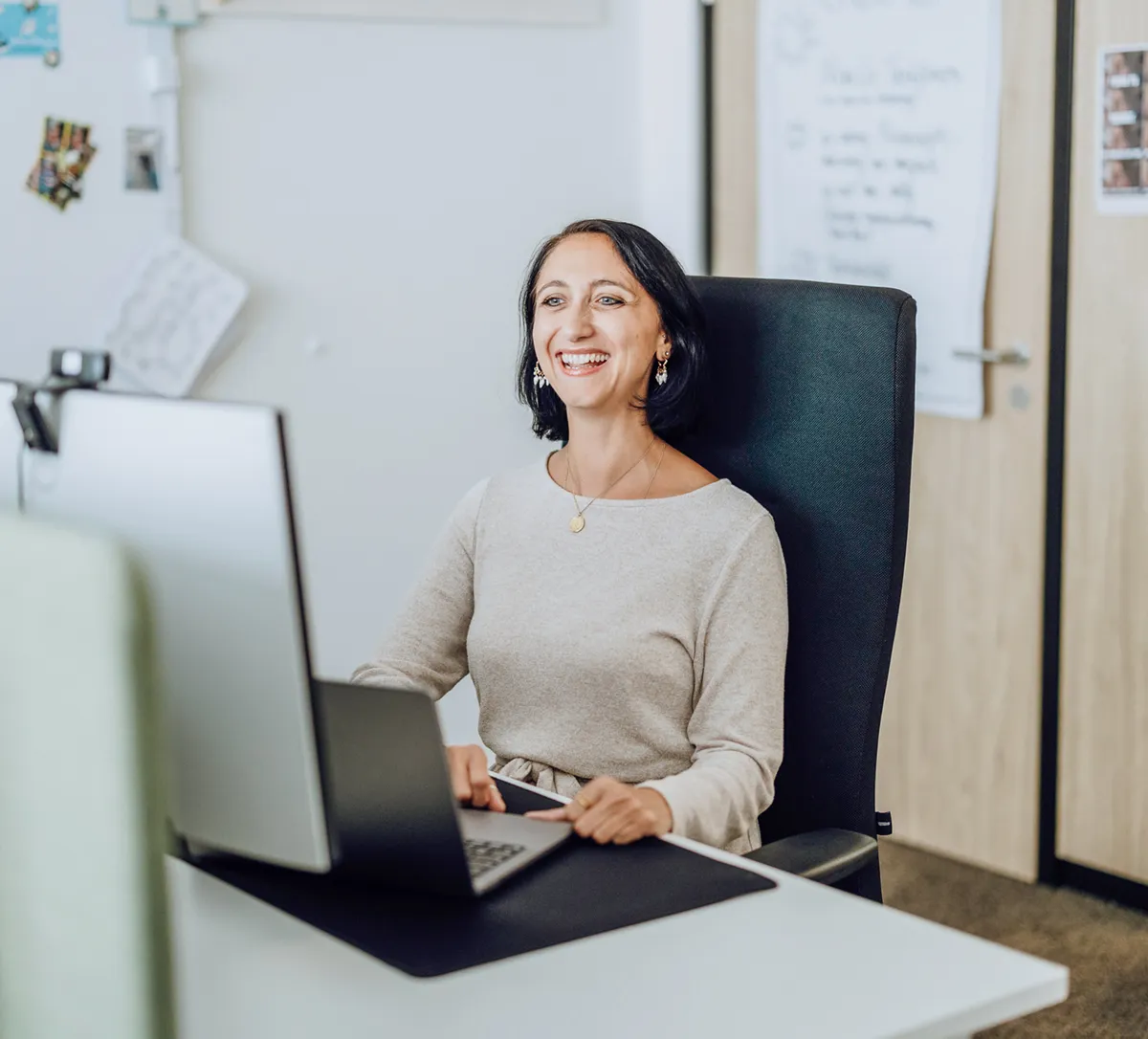 Woman smiling while working at a desk with a laptop and dual monitors in an office setting.